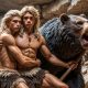 Two muscular male cavemen with long blonde hair embrace protectively in front of a roaring bear. One holds a spear as they stand in a rocky cave setting, while other cavemen appear in the background.