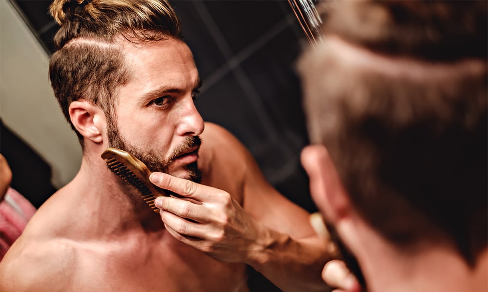 Hipster man focusing on combing beard in front of a mirror