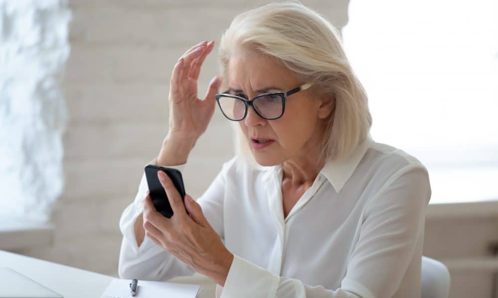 Confused senior businesswoman sit at office desk hold cellphone
