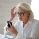 Confused senior businesswoman sit at office desk hold cellphone