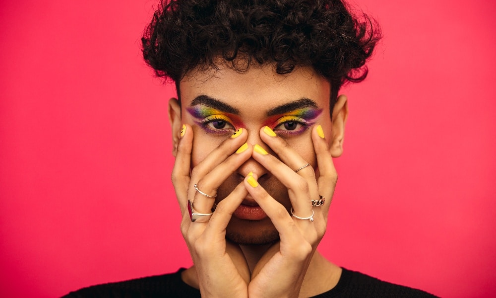 Gender fluid person wearing rainbow colored eye shadow and smiley face on fingernail. 