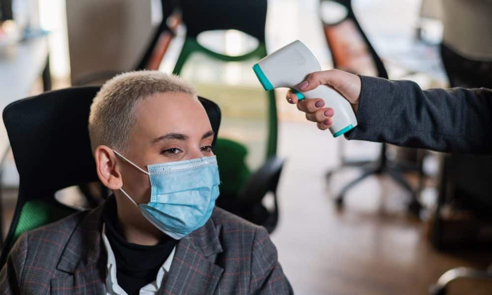 Business woman in medical mask has body temperature measured with electronic thermometer at workplace in office.