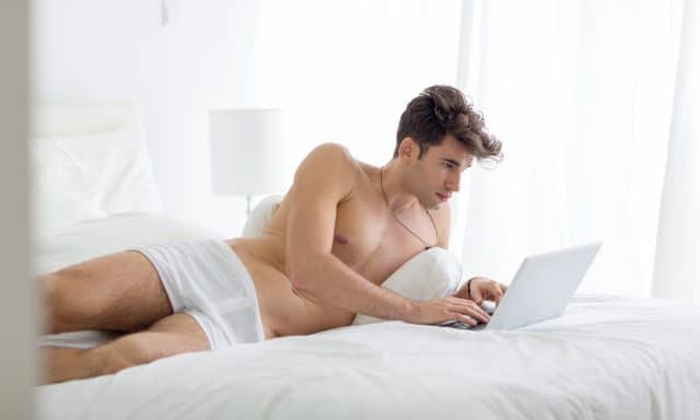 Young shirtless man working on a laptop computer lying in bed during the day