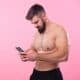 Shirtless strong bodybuilder talking on his cell, using a smartphone, pink studio background