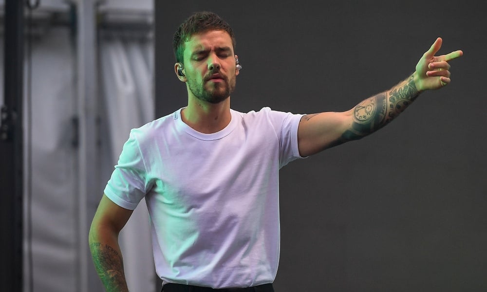 Liam Payne, former One Direction singer, performs on stage wearing a white t-shirt, with his left arm raised and eyes closed. His arm displays several visible tattoos, including a clock on his forearm. The background is a neutral concert setting.