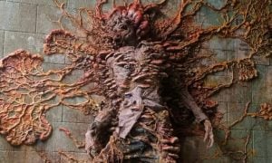 This image from The Last of Us depicts a chilling scene of a human infected by the "Cordyceps" fungus. The fungal growth has overtaken the host, transforming them into a grotesque and surreal figure. The body appears to be fused to a wall, with vibrant fungal tendrils and spores spreading outward, showcasing the invasive nature of the infection. This representation highlights the post-apocalyptic horror and the terrifying consequences of a parasitic fungus controlling its host in the game's world.