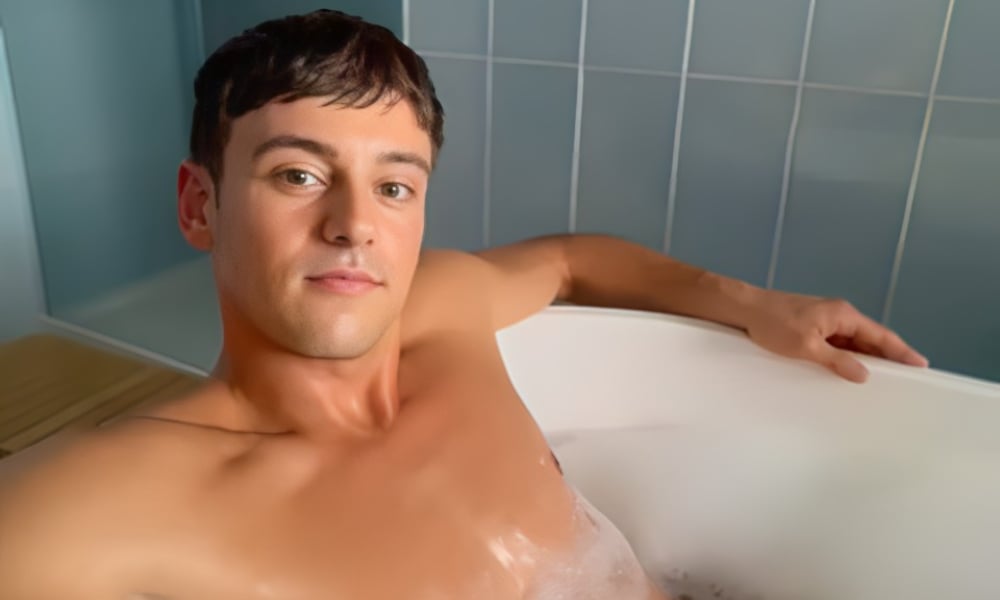 Tom Daley lounging in a bubble-filled bathtub, with a relaxed expression on his face, his right arm resting on the tub's edge.